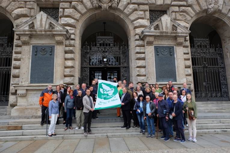 Eine große Gruppe von Menschen unterschiedlichen Geschlechts und Alters hält die grün-weiße Mayors-for-Peace-Flagge vor dem Rathaus in Leipzig in die Kamera.